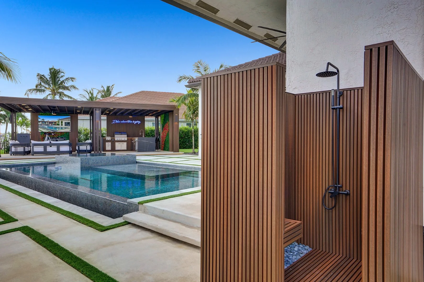 Poolside terrace with a slatted timber outdoor shower in the foreground and a covered lounge and outdoor kitchen beyond