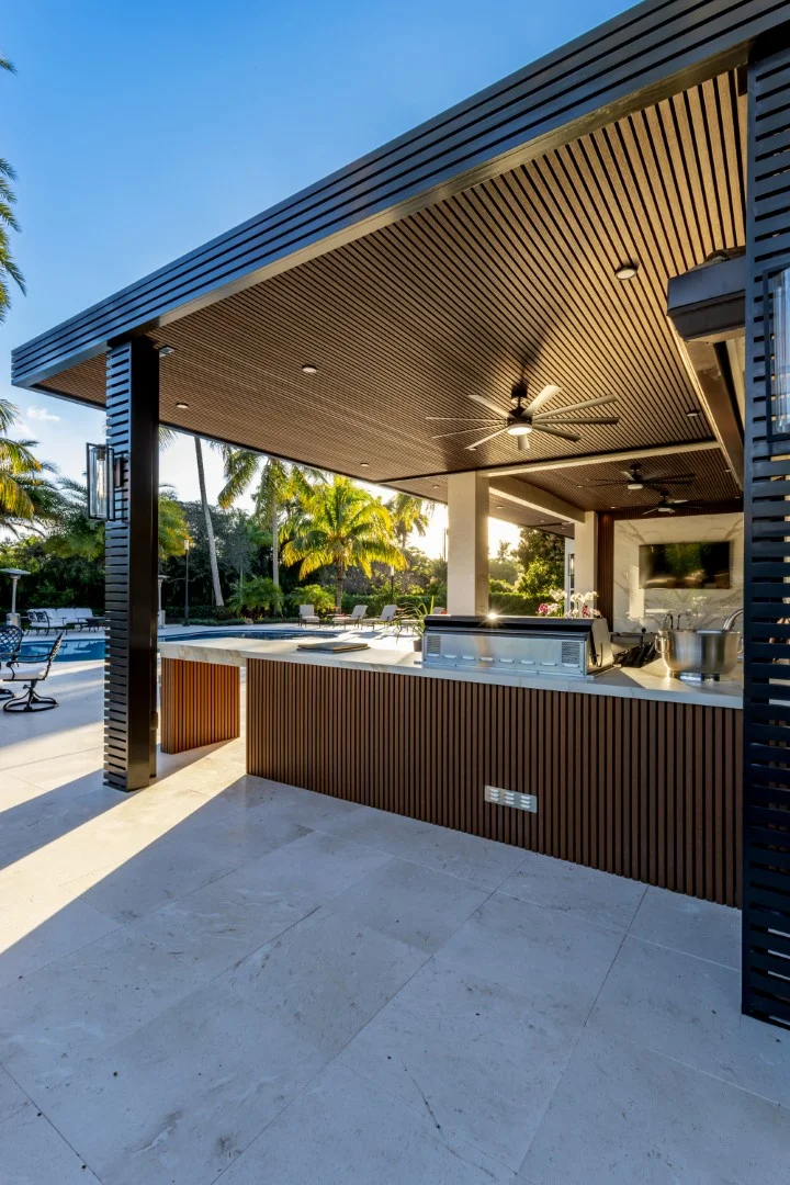 Outdoor kitchen beneath a slatted timber pergola ceiling, flanked by dark privacy screens beside a pool